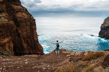 Adventurous man standing on rocky cliff edge, arms outstretched, overlooking vast ocean waves and dramatic coastal landscape, embodying freedom and exploration in nature's beauty