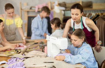 Hardworking preteen schoolgirl intently stitching at sewing machine during group lesson, fully absorbed in task while peers working on fabric and designs with help from female instructor..