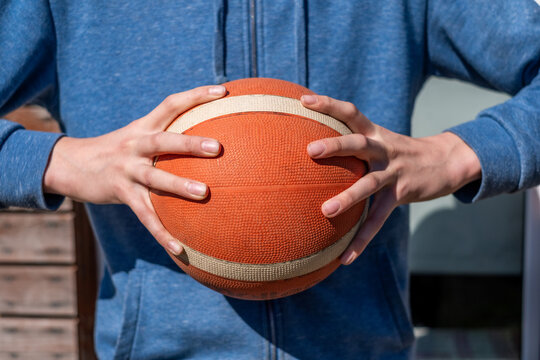 Young male athlete in blue hoodie holds a basketball with both hands, showcasing grip and focus, ready for action on outdoor court with blurred background