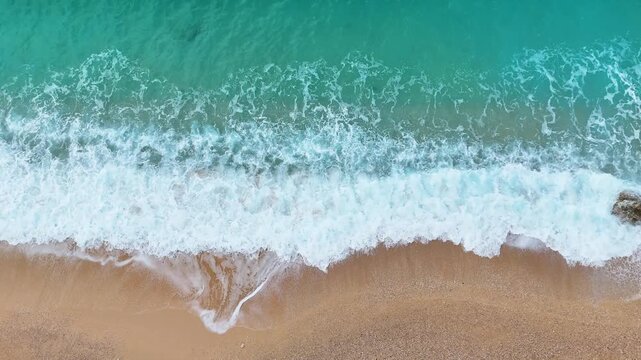 Stunning top-down aerial drone view capturing dynamic turquoise ocean waves and detailed white sea foam crashing onto a pristine sandy beach shoreline. A perfect tropical scene.