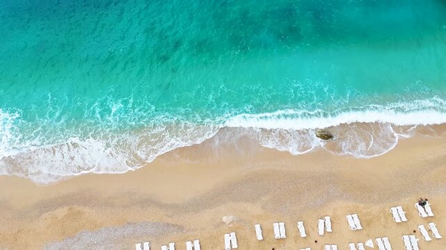 Cinematic top-down drone shot capturing a breaking turquoise ocean wave onto a golden sand beach, creating beautiful white foam patterns. Serene and picturesque tropical coastal landscape view.