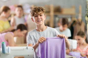 Teen boy student of school of young fashion designers carefully examines fabric, evaluates product and work performing. Training sessions for students of sewing courses...