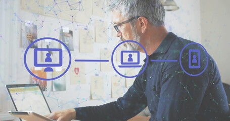 Working senior man wearing glasses, dark sweater using laptop, tablet in home office, network icons