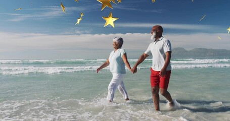 Walking mature couple in beach clothes holding hands through surf at shoreline, gold stars