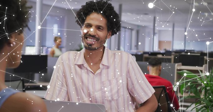 Smiling man wearing pink striped short-sleeve shirt talking at open-plan office, laptop and nodes - Powered by Adobe