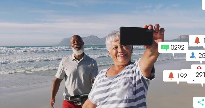 Smiling senior woman in striped top taking selfie at beach with phone, man by bike, notifications