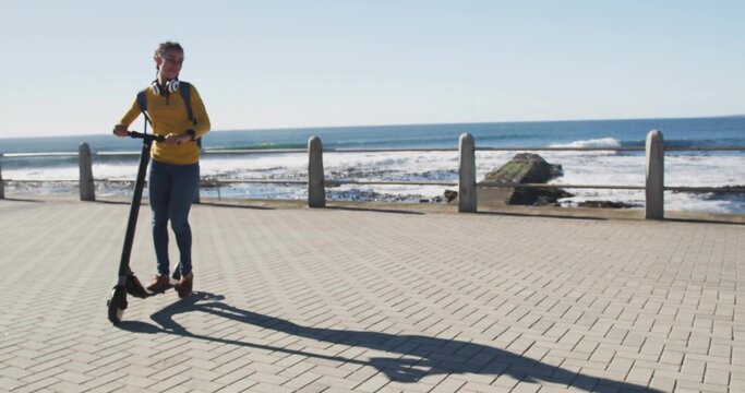 Riding woman wearing yellow top on seafront, holding e-scooter with headphones backpack, copy space - Powered by Adobe