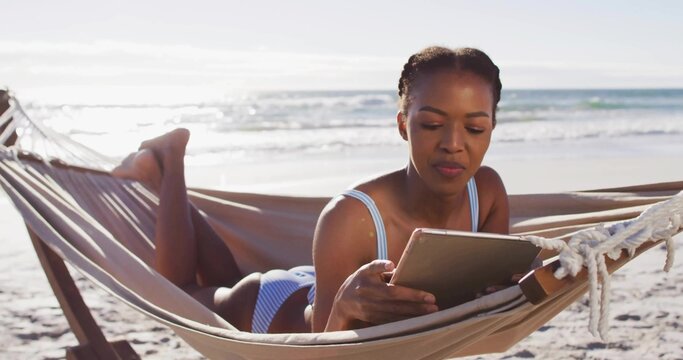 Reclining woman reading tablet on sandy ocean beach, wearing light blue striped swimsuit on hammock
