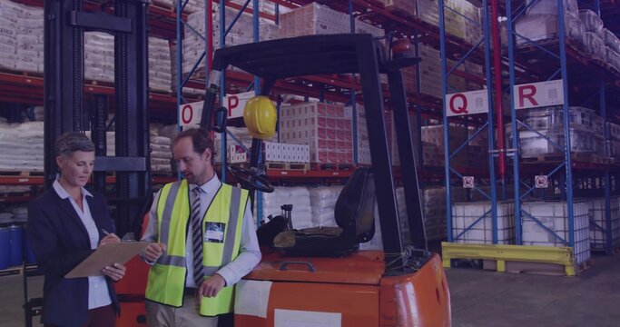 Checking woman in blazer and man in vest counting pallets in warehouse with orange forklift, helmet