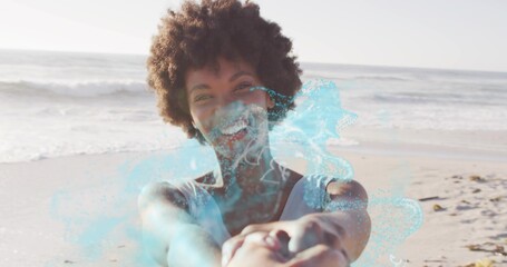 Smiling woman in sleeveless top holding partner's hands at beach shore, breaking waves, blue splash