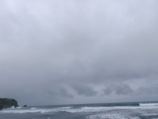 Wide view of a dark ocean under a heavy gray cloudy sky. Dramatic storm clouds hanging low above the vast open sea. Overcast horizon showing dark waves and a rocky coastal island.