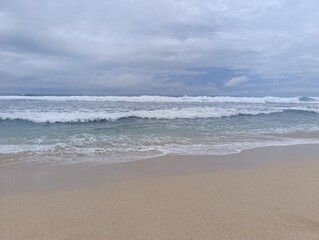 Large blue ocean wave cresting and breaking on the wide beach. Dynamic motion of white sea foam over clear blue water on the shore. Powerful coastal wave washing onto the light brown beach sand.