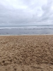 Wide empty beach scene of brown sand meeting the calm ocean waves. Calm sea water and overcast sky . Tranquil beach landscape on a cloudy day with footprints in the sand.