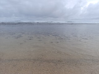 Calm shallow water covering the exposed reef during a low tide. Ocean water receding over the brown textured sand and rock bed. Natural abstract reflections of the cloudy sky on the low water surface.