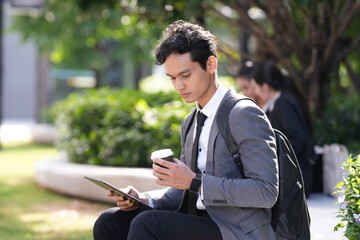 Young Asian businessman with backpack sitting in green city park, holding coffee and using digital tablet for remote work during break.