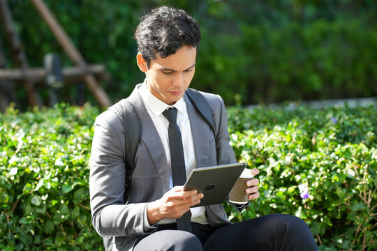 Young Asian businessman with backpack sitting in green city park, holding coffee and using digital tablet for remote work during break.