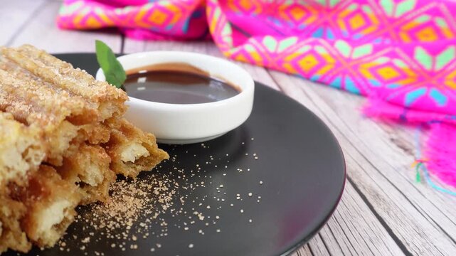 Pan to a plate of churros with cinnamon, sugar, and chocolate on a wooden table