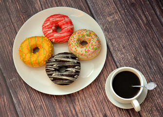 Four fresh doughnuts with colorful glaze and toppings and a cup of hot black coffee on a wooden table.