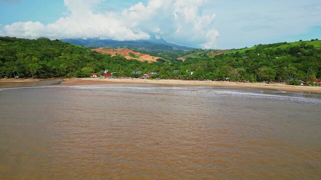 Rising aerial showing sandy shoreline with brown water waves rolling onto the coast, framed by lush hills and a small town along the coastline in Mariveles, Bataan.