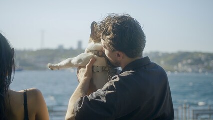 Man Holding Dog by the Water with Woman in Background