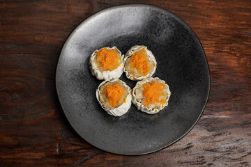Four steamed dimsum with grated carrot topping placed at the center of a dark plate, shot in a centered composition over a rustic wooden background.
