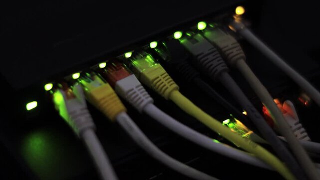 A close-up shot of colorful Ethernet cables plugged into a network switch, highlighted by bright green connection lights in a dark environment.