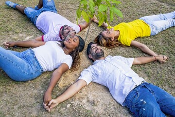 Diverse friends lying in circle holding hands around a planted tree