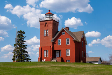 The Two Harbors Lighthouse and attached home, near the shore of Lake Superior in Minnesota, on a sunlit spring day with blue shy and fluffy white clouds.