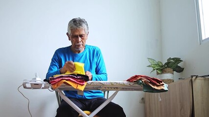 Elderly asian man carefully folds colorful laundry on ironing board