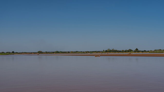 A calm red-brown African river. The pirogue boat floats in the distance. Green vegetation on a sandy beach. Clear blue sky. Madagascar. Tsiribihina river.
