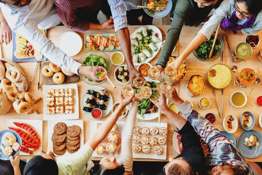 A diverse group of people, including men and women of various ethnicities, joyfully sharing a feast. The table is filled with delicious food, showcasing a vibrant and communal dining experience. - Powered by Adobe