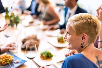 A group of people enjoying a meal together at a restaurant. Focus on a woman with short blonde hair, smiling, with wine and food on the table. Social dining scene. Business lunch for business people.