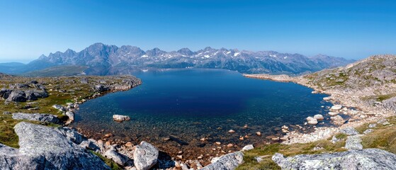 A panoramic view of a deep blue alpine lake surrounded by rocky terrain and distant, snow-capped mountains under a bright, clear blue sky.