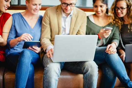 A diverse group of professionals, including women and men of various ethnicities, collaborate using laptops and smartphones. This teamwork showcases modern technology in a casual setting.