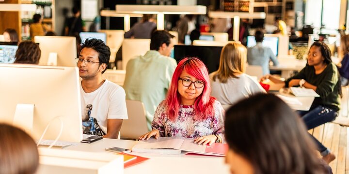 Diverse group of people working in a library. Individuals focused on computers, collaborating in a busy workspace. Office environment with teamwork and technology. Diverse people working at library.
