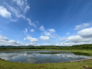 Wide reservoir view with bright blue sky, clouds, and lush green hills surrounding calm water.