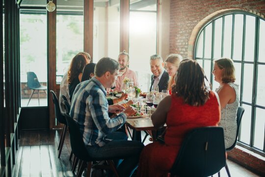 Group of people enjoying a meal together at a dining table in a cozy, sunlit room. Laughter and conversation fill the air as they share food and drinks. Celebrations, Communal dining.