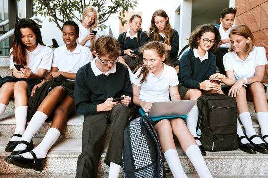 Group of diverse students in school uniforms sitting on steps. Students using laptops and phones. Boys and girls in uniforms. School uniforms and technology. Diverse teenage students at school.