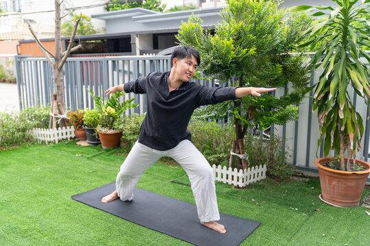 Young man practicing yoga outdoors on black mat in garden, stretching arms with focused expression, surrounded by green plants and trees in residential area