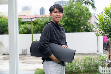 Young man holding black yoga mat outdoors in casual clothing with greenery and urban background, relaxed and ready for exercise or meditation