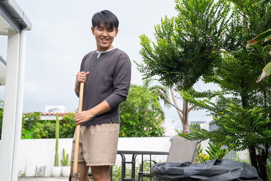 Young man cleaning outdoor garden area with broom, smiling and enjoying home cleaning on bright day with green plants and trees around - Powered by Adobe