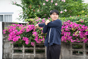 Man stretching arm outdoors in front of pink flowers and green bushes, wearing navy jacket and black pants, preparing for exercise in casual setting