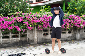 Young man stretching arms outdoors near dumbbell weights on pavement with vibrant pink flowers and green foliage in background, showing fitness and relaxation in natural setting