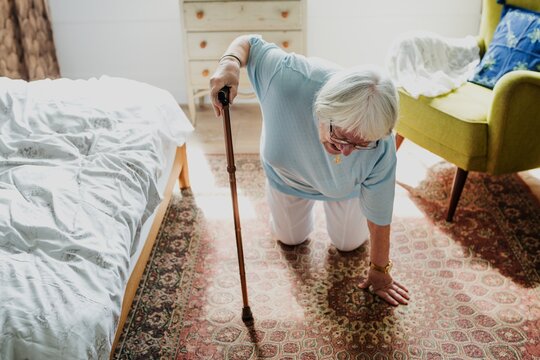 Elderly woman with cane kneeling on carpet. Senior woman, cane, carpet. Older adult, cane, carpet. Elderly female, cane, carpet. Home setting, cane, carpet. Old woman falling to the floor in care home