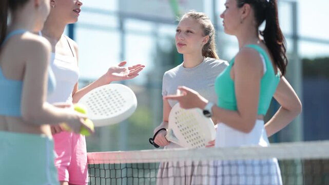 Athletic women stand near the net on the court, hold rackets and chat in between playing paddle tennis. High quality 4k footage