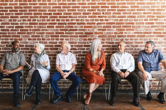 Group of diverse seniors sitting on chairs against a brick wall, engaged in conversation. Elderly people, casual attire, diverse backgrounds, friendly interaction. Group of people sitting together.