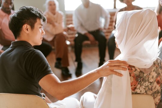 Asian man comforting muslim woman in hijab during support group meeting, diverse people. Support group of diverse people, Asian man and muslim woman share supportive moment in diverse support group - Powered by Adobe
