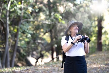 Smiling senior woman enjoying nature with binoculars. Senior woman with binoculars, wearing a hat, exploring nature with binoculars. Senior couple enjoying outdoors adventure.