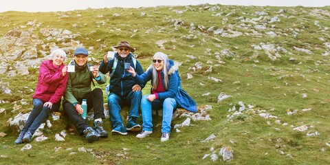 Group of four elderly friends sitting outdoors on a grassy hill, smiling and holding drinks. Casual attire, enjoying nature trip and friendship. Happy retirement. Traveling with elderly friends