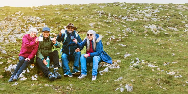 Group of four elderly friends sitting outdoors on a grassy hill, smiling and holding drinks. Casual attire, enjoying nature trip and friendship. Happy retirement. Traveling with elderly friends